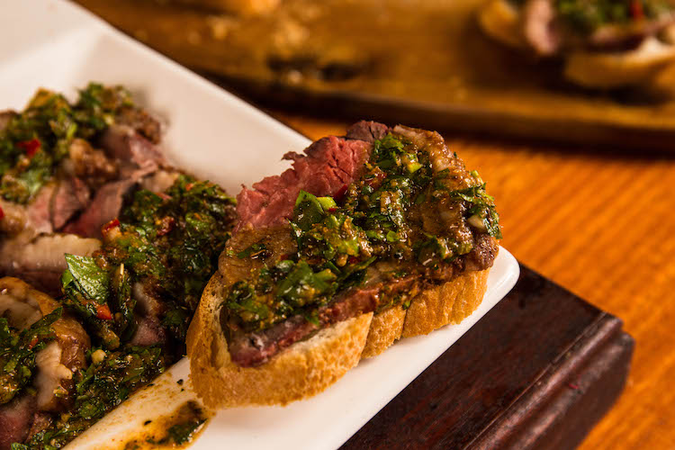 a piece of baguette with a slice of cooked picanha steak and chimichurri sauce
