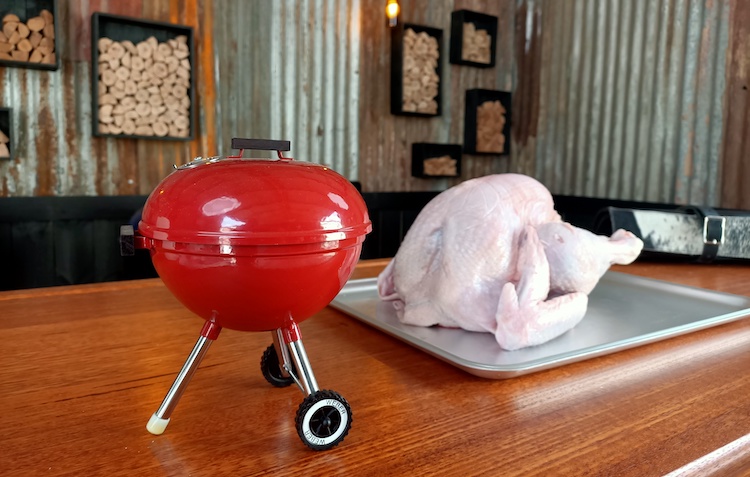uncooked whole turkey on a wooden table with a toy weber kettle grill beside it