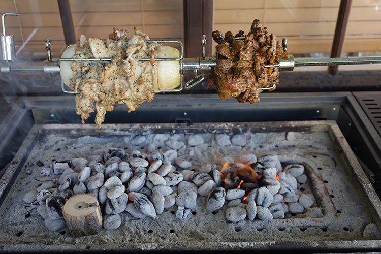 sliced chicken and lamb shoulder cooking on a rotisserie rod with charcoal burning underneath