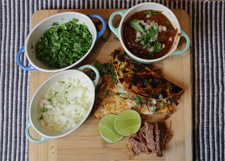 birria taco, consomme, chopped onions, cilantro and half of lime on a wooden board