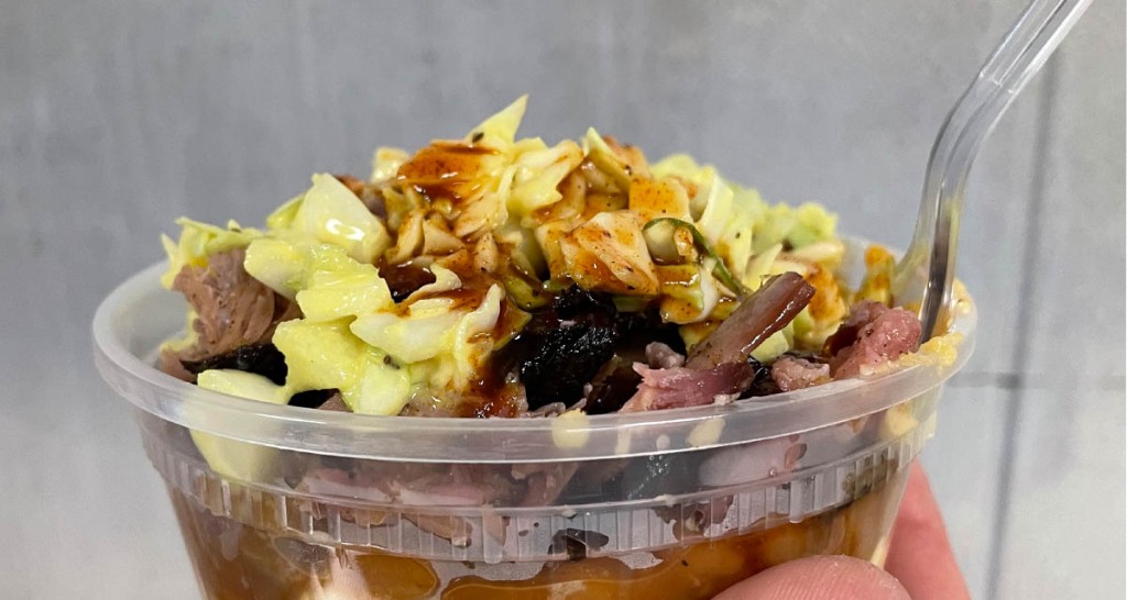 clear container layered with pork, beans, mac n cheese and bbq sauce with a spoon in it