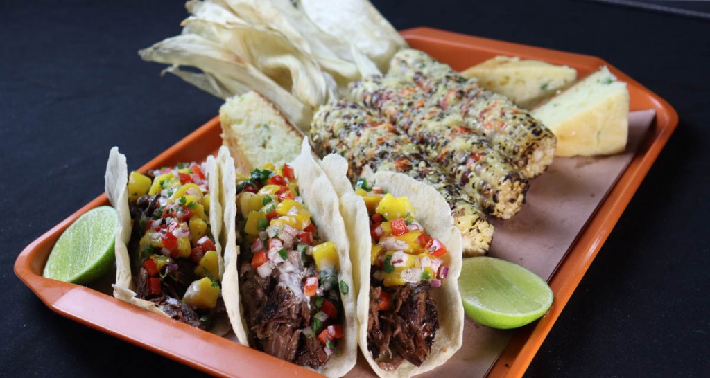 smoked beef shank tacos on a tray with grilled corn cobs, jalapeno bread and a lime cheek