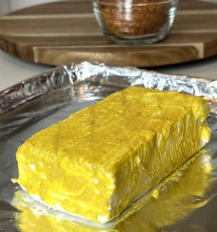 a block of cream cheese covered in yellow mustard on a sliver tray