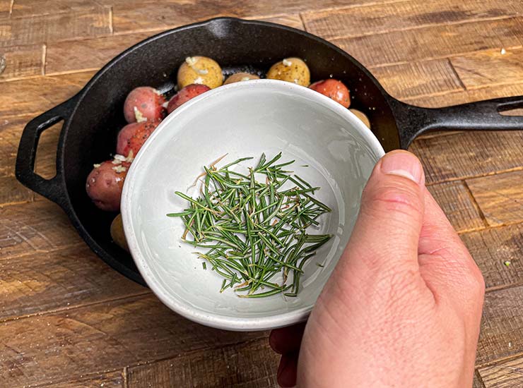 rosemary leaves in a white bowl and a cast icon skilled with baby potatoes on the background