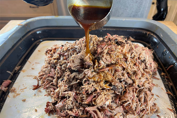 shredded pork butt in tray with juices being poured oover it