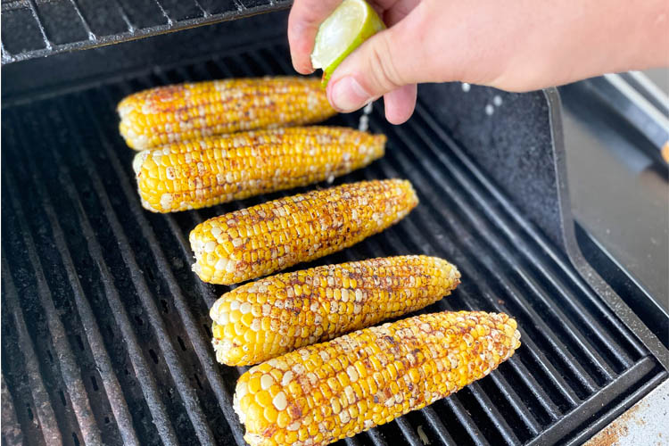 five corn cobs on the grill with a hand squeezing lime