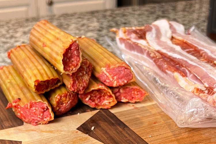 stuffed raw manacotti on a wooden chopping board with bacon strips beside