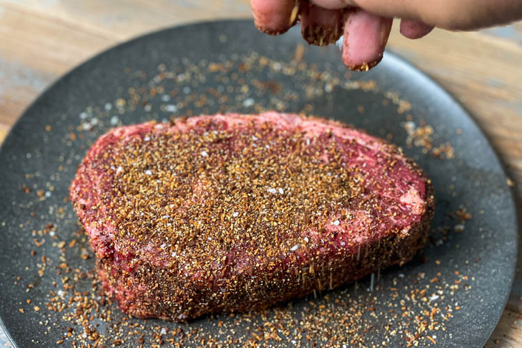 a hand sprinkling rub on a ribeye steak on a black plate