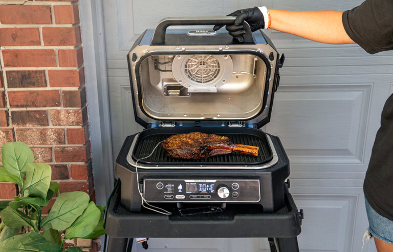 A cooked tomahawk steak rests on the grill grate of a Ninja Woodfire Pro XL, with the lid open to reveal the interior heating fan. A hand wearing a black glove holds the lid open, while the grill sits on a black cart in front of a garage door and red brick wall.