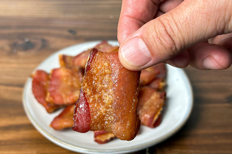 fingers holding a bacon wrapped cracker, with a plate of them in the background