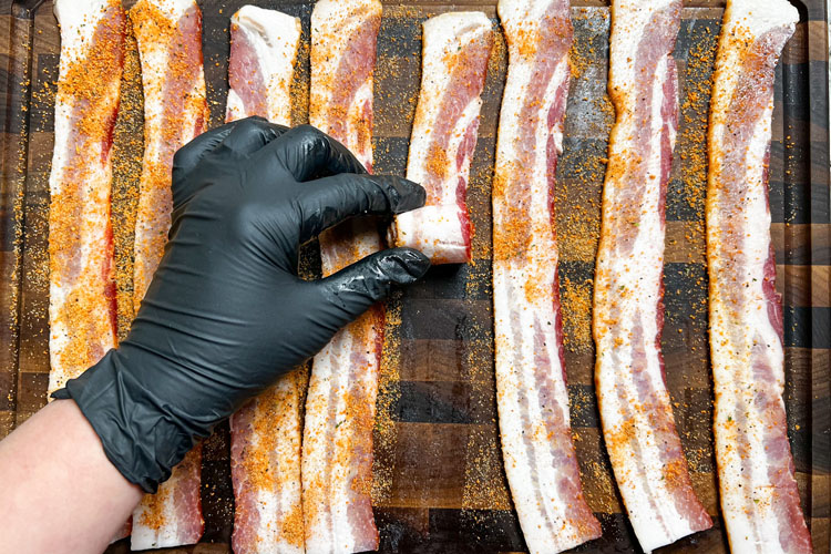seasoned bacon laid on a wood board and a black gloved hand rolling a rasher up