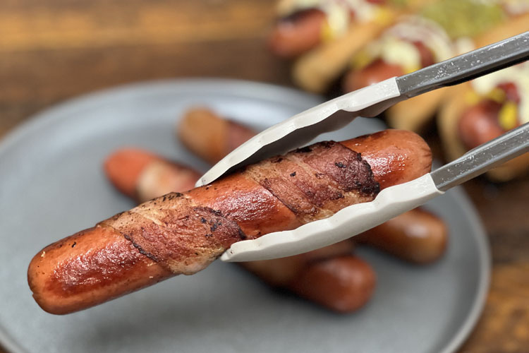 a bacon-wrapped hot dog being held over a plate by some tongs