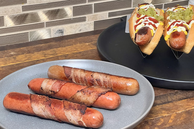 three bacon-wrapped hot dogs on a grey plate 