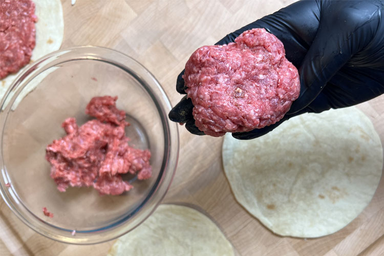 a ball of ground beef being held by a black gloved hand, with mince in a bowl in the background