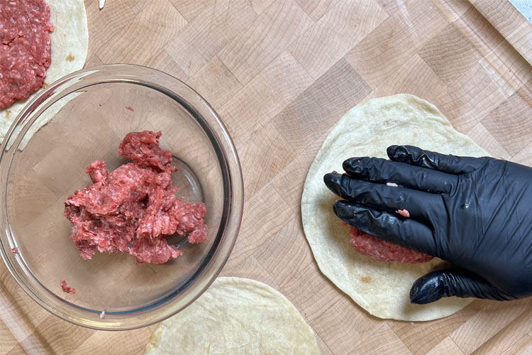 ground beef beef being squished onto a flour tortilla by a black gloved hand