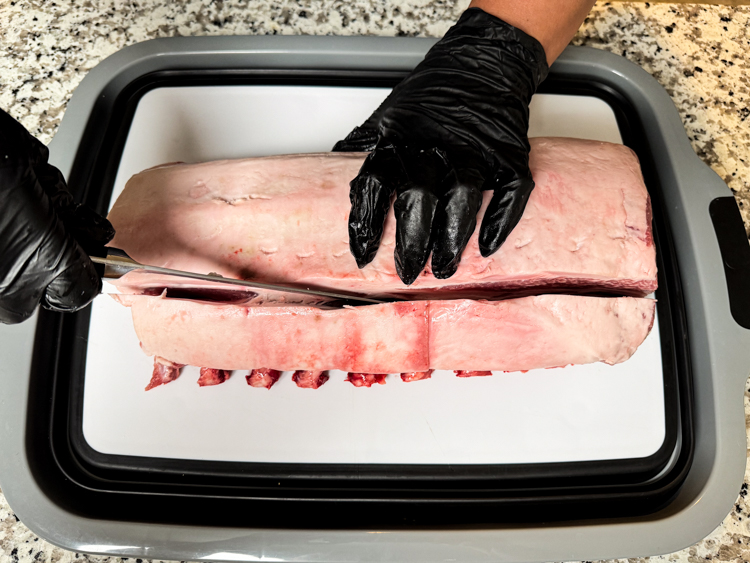 a black gloved hand holing a bone in rack of pork slicing along the top with a knife