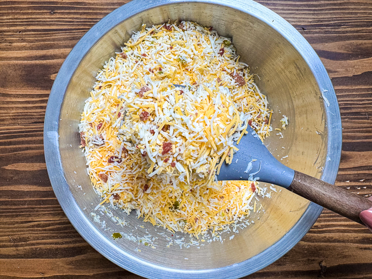shredded cheese, bacon bits and jalapenos in a metal bowl with a rubber spoon in it