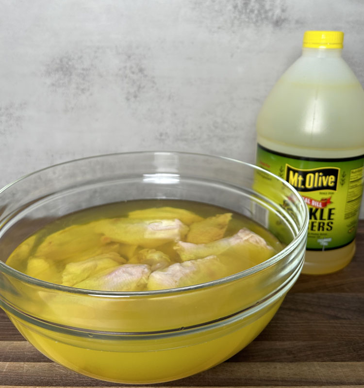 a glass bowl with wings marinating in pickle juice and a bottle of pickle juice in the background