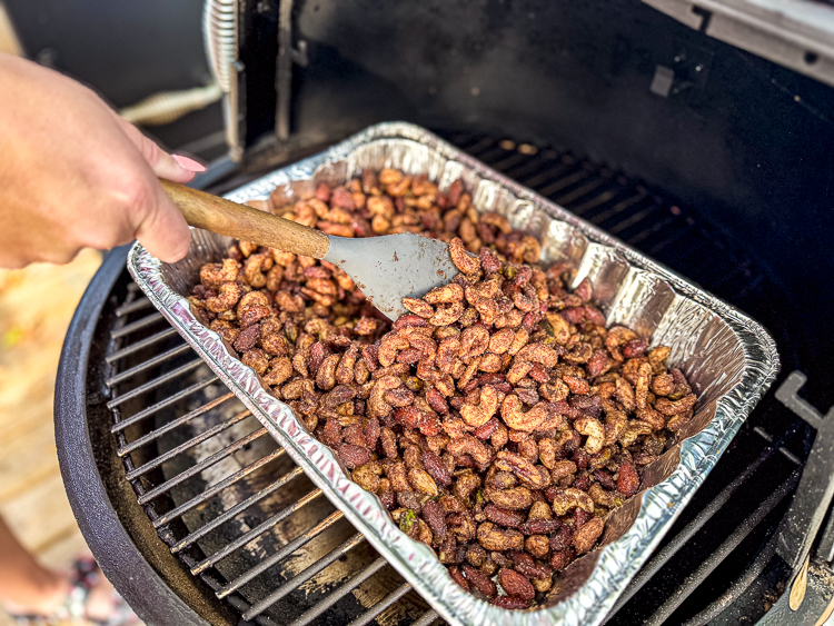 a wooden spoon stirring a tray of candied nuts on the smoker