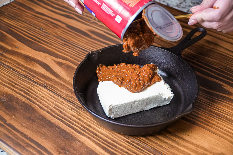 pouring a can of chili over cream cheese in a skillet