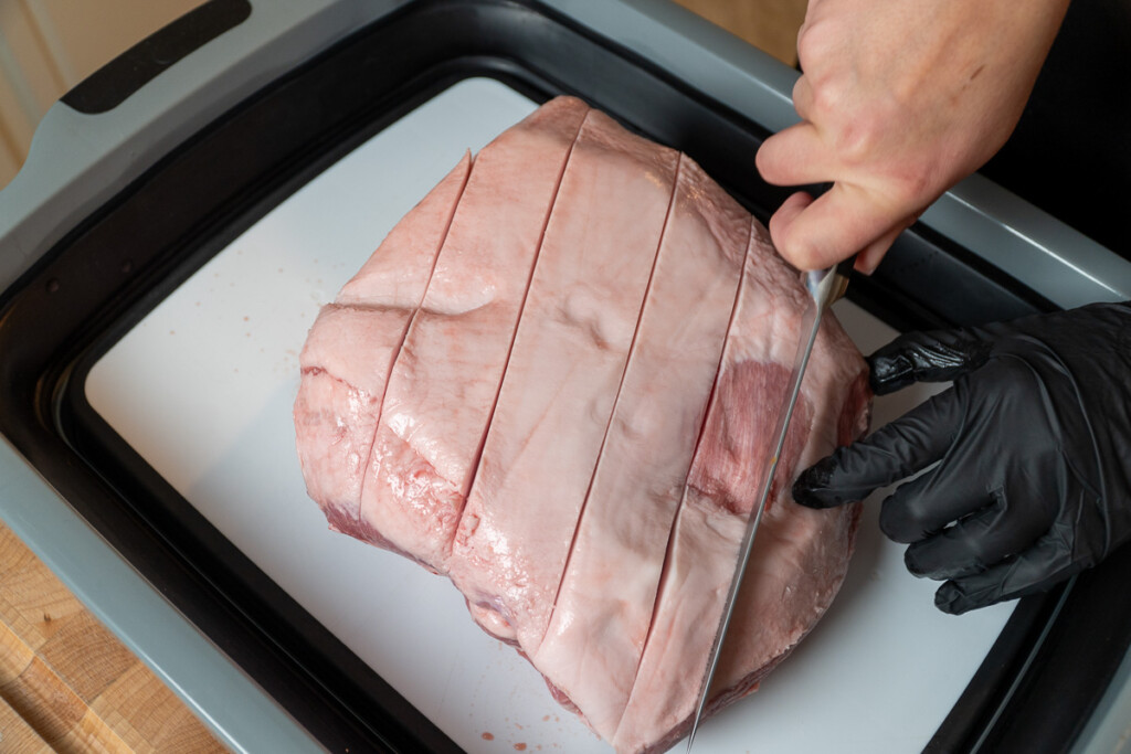 a hand with a knife slicing the skin of a piece of pork butt