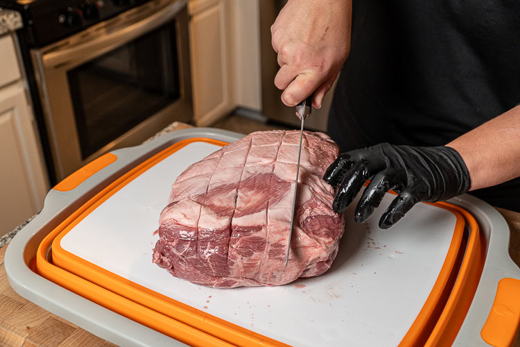 hand holding knife making cuts into the fatcap of a raw piece of pork butt