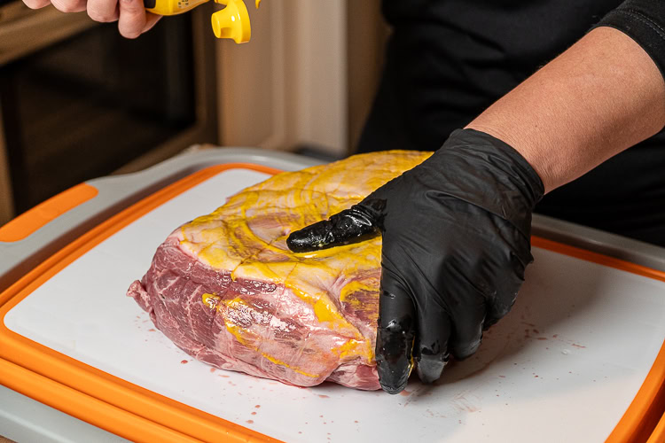 black gloved hand rubbing mustard onto the surface of a piece of pork but