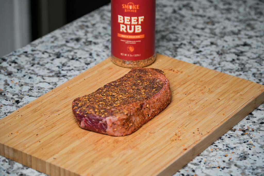 a seasoned raw steak and bottle of smoke kitchen rub on a wooden chopping board