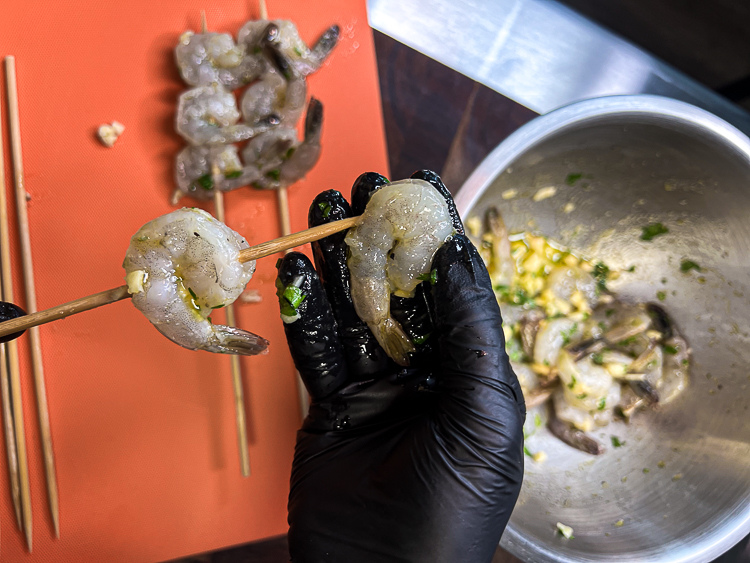 a black gloved hand threading shrimp on to a babmoo skewer