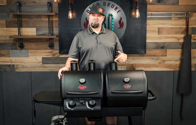 Man standing behind and pointing at the Char-Griller Dual Fuel 5030 gas and charcoal combo grill in a workshop setting, with a wooden wall and shelves in the background. Ask ChatGPT