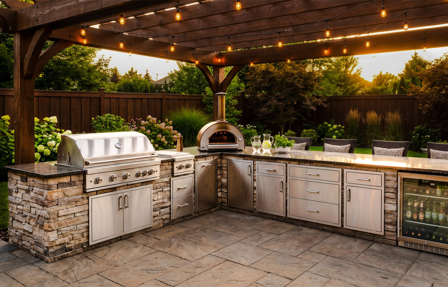 A fully equipped outdoor kitchen with built-in gas grill, wood-fired pizza oven, stainless steel storage drawers, and beverage fridge under a wooden pergola with string lights.